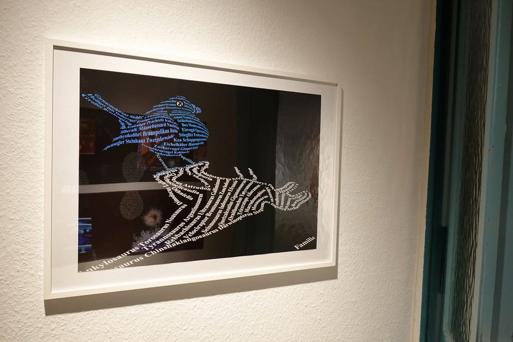 Gerahmte grafische Arbeit an einer Galeriewand: Zwei dinosaurierartige Figuren, zusammengesetzt aus vielen Wörtern, stehen einander gegenüber; eine ist blau, die andere weiß auf dunklem Hintergrund.